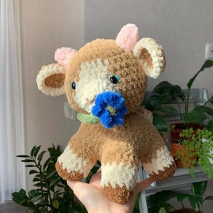 Op de afbeelding: Een handgemaakte gehaakte koeienknuffel in bruin, cr&egrave;me en wit. De koe heeft blauwe ogen, roze oren en een blauwe bloem in haar mond. De knuffel is zacht en knuffelig, ideaal als kinderspeelgoed of voor decoratie.