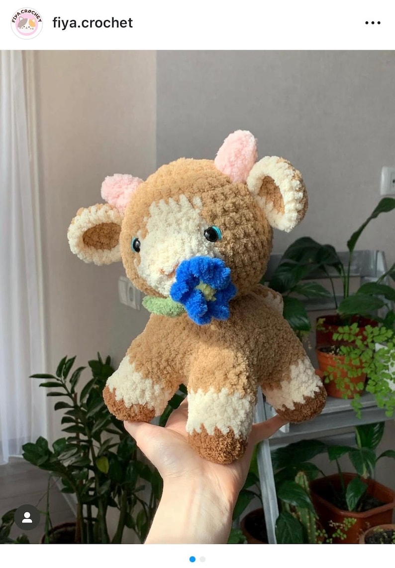 Op de afbeelding: Een handgemaakte gehaakte koeienknuffel in bruin, cr&egrave;me en wit. De koe heeft blauwe ogen, roze oren en een blauwe bloem in haar mond. De knuffel is zacht en knuffelig, ideaal als kinderspeelgoed of voor decoratie.