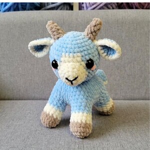 Op de afbeelding: Een handgemaakte gehaakte geit in lichtblauw, wit en bruin. Het amigurumi-dier heeft grote zwarte ogen en kleine hoorns. De tekst "betsymadecrochet" is zichtbaar. Het speelgoed is handgemaakt en staat op vier poten.