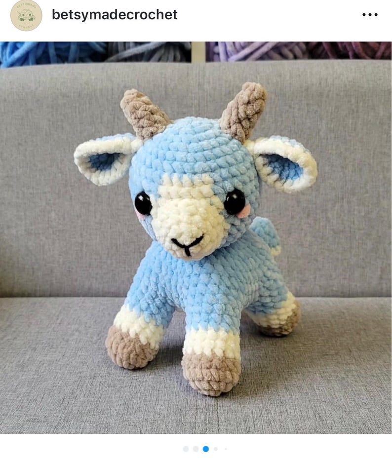 Op de afbeelding: Een handgemaakte gehaakte geit in lichtblauw, wit en bruin. Het amigurumi-dier heeft grote zwarte ogen en kleine hoorns. De tekst "betsymadecrochet" is zichtbaar. Het speelgoed is handgemaakt en staat op vier poten.