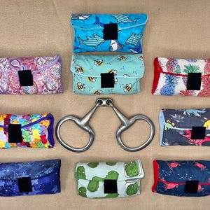 May include: A selection of patterned fabric pouches featuring designs such as sharks, bees, pineapples, and dinosaurs. Each pouch has a black square closure. A silver metal horse bit is in the center.