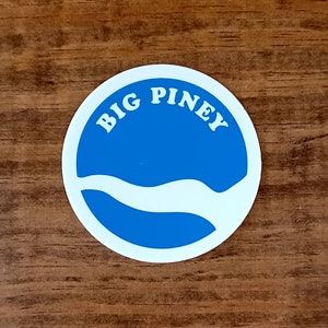 May include: A white circle sticker with a blue background and a white wave design. The text "BIG PINEY" is written in white letters around the circle.