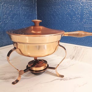 May include: A copper-colored chafing dish with a lid and wooden handle. The dish sits on a stand with curved legs and a burner below. The lid has a decorative pattern.