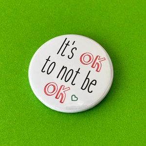 May include: A white badge with the words "It's OK to not be OK" in black and red. A small green heart is at the bottom. The badge is set against a green backdrop.