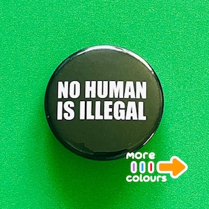 Könnte beinhalten: Ein runder, dunkelgrüner Button mit dem weißen Text "NO HUMAN IS ILLEGAL". Der Button hat eine glänzende Oberfläche und befindet sich vor einem hellgrünen Hintergrund. Der Button hat auch die Worte "more colours" und einen Pfeil.