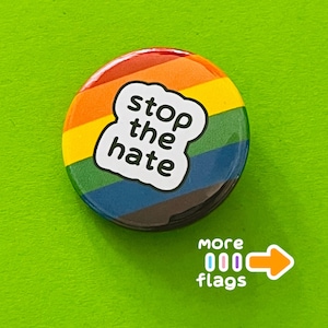 May include: A round button with a rainbow design and the words "stop the hate" in a white cloud-shaped text box. The button is on a green surface, with an arrow and the words "more flags".