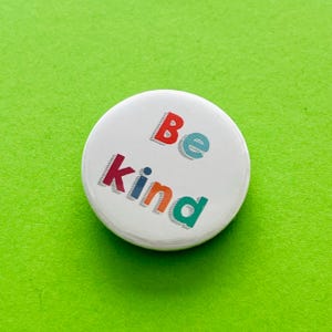 May include: White button with the words "Be kind" in colourful, outlined letters.