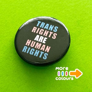 May include: A round black pin-back button featuring the text TRANS RIGHTS ARE HUMAN RIGHTS in light blue, pink, and white lettering. Below the button, the text more colours is printed in white next to an orange arrow pointing to the right.
