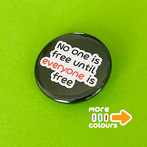 Könnte beinhalten: Ein runder schwarzer Ansteckbutton mit weißem Text, der No one is free until everyone is free lautet, wobei das Wort everyone in Rot hervorgehoben ist. Unter dem Button steht der Text more colours mit drei kleinen Ovalen und einem Pfeil.