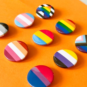 May include: A selection of circular enamel badges displaying LGBTQ+ pride flags. The badges feature striped and geometric designs in rainbow, pink, blue, yellow, and black. They are arranged on an orange surface.