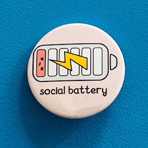 May include: A circular, pale pink badge with a black border. It displays a battery icon with a yellow lightning bolt and the words "social battery" in black. The battery graphic indicates a low charge.