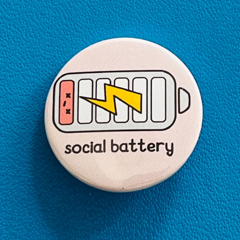 May include: A circular, pale pink badge with a black border. It displays a battery icon with a yellow lightning bolt and the words "social battery" in black. The battery graphic indicates a low charge.