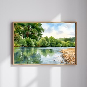 Può includere: Un dipinto ad acquerello rettangolare in una cornice di legno che raffigura un fiume calmo che scorre accanto a una fitta foresta di alberi verdi. La scena presenta una riva di ciottoli sulla destra e riflessi degli alberi sull'acqua.