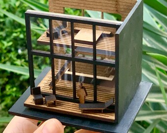 May include: A miniature wooden model of a modern house with a black frame and glass windows. The house has two levels and a small balcony. The model is sitting on a black base.