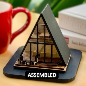 May include: A wooden model of a modern A-frame house with black trim and glass windows. The model is assembled and sitting on a black base with the word "ASSEMBLED" printed on it.