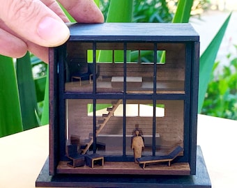 May include: A miniature wooden model of a modern house with a black frame and glass windows. The house has a staircase, a figure standing inside, and furniture. The model is sitting on a black base.