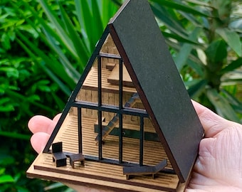 May include: A miniature wooden model of an A-frame house with a black roof and large windows. The model is sitting on a wooden base and has a small deck with furniture.