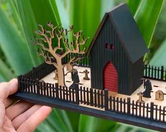 Op de afbeelding: Miniatuur houten diorama met Halloween-thema met een zwarte kerk met een rode deur, een begraafplaats met grafstenen en een kale boom. De scène is omgeven door een zwart hek, wat een spookachtige sfeer creëert.