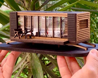 May include: A miniature wooden model of a small house with a glass front and a wooden deck. The model is painted brown and has a black roof. The model is sitting on a black base.