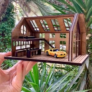May include: A miniature wooden house model with a two-story design, featuring a loft, staircase, and large windows. A yellow toy car with flame decals is parked inside. The model is held against a backdrop of green foliage.