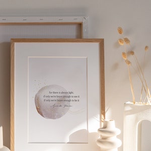 May include: A framed print with a watercolor-style illustration of a circle with a quote inside. The quote reads "For there is always light, if only we're brave enough to see it. If only we're brave enough to be it." The print is on a white shelf with two white ceramic vases and dried flowers.