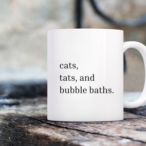 May include: White ceramic mug with black text that reads "cats, tats, and bubble baths."