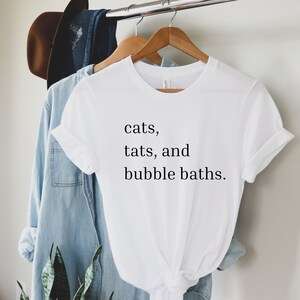 May include: White t-shirt with the text "cats, tats, and bubble baths." hanging on a wooden hanger.