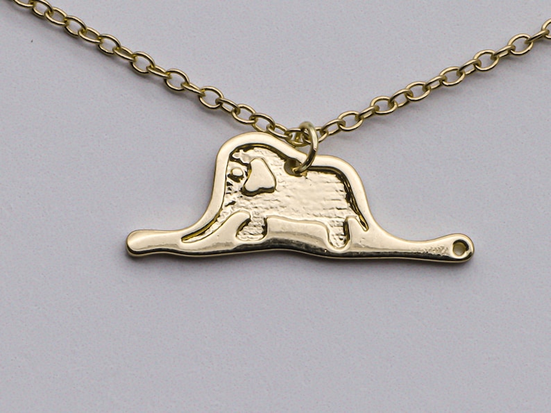 May include: A gold necklace with a pendant shaped like an elephant standing on a thin line. The elephant is detailed with textured lines and a small heart shape on its chest.