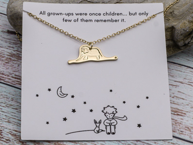 May include: Gold necklace with a charm of the Little Prince and the fox standing on a planet with a crescent moon and stars in the background. The card reads "All grown-ups were once children... but only few of them remember it."