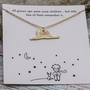 May include: Gold necklace with a charm of a small prince and a fox, and a quote from The Little Prince: "All grown-ups were once children... but only few of them remember it."