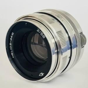 May include: A silver Helios-44M 2/58 lens for a 35mm camera. The lens has a black aperture ring and a silver focusing ring.