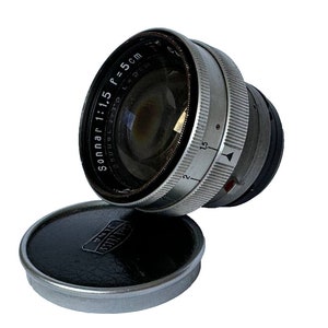May include: A silver and black camera lens with the text "Sonnar 1:1.5 f=5 cm" printed on the side. The lens is attached to a black lens cap.