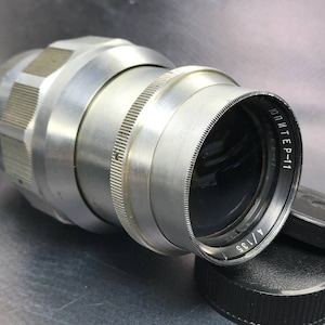 Può includere: Un obiettivo argentato con la scritta "Юпитер-11" stampata sul lato. L'obiettivo ha un tappo dell'obiettivo nero e un parasole nero.