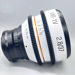 May include: A professional camera lens, the MIR-1V 2.8/37, is white with black and orange accents. The lens has multiple adjustment rings with markings for aperture and focus. The lens is designed for 8mm-52mm.