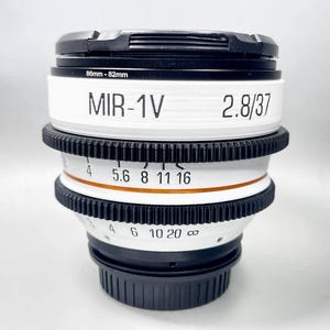May include: A white and black MIR-1V 2.8/37 camera lens with a black lens cap. The lens has a focus ring with numbers and markings. The lens is 37mm and has a filter size of 86mm-82mm.