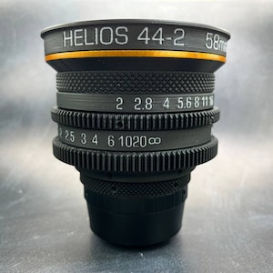 May include: A black Helios 44-2 58mm lens with a gold ring around the front. The lens has a depth of field scale with markings for 2, 2.8, 4, 5.6, 8, 11, 16, 22, 25, 3, 4, 6, 10, 20, and infinity.