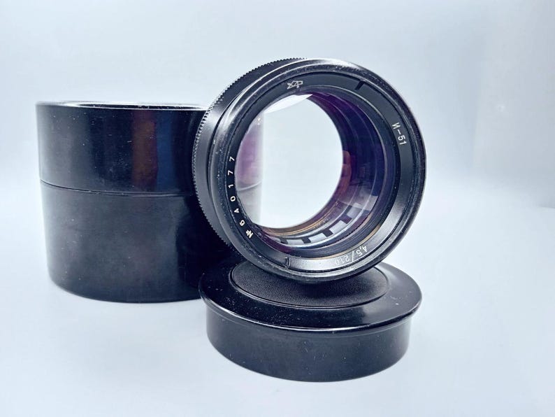 May include: A black camera lens with a silver ring and markings. The lens is sitting on a black lens cap.