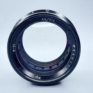 May include: A black camera lens with a silver ring and markings. The lens has a silver inscription that reads "M-51" and "4,5/210". The lens also has a silver inscription that reads "N 640171".