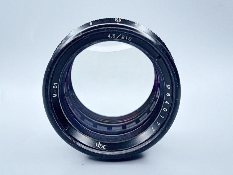 May include: A black camera lens with a silver ring and markings. The lens has a silver inscription that reads "M-51" and "4,5/210". The lens also has a silver inscription that reads "N 640171".