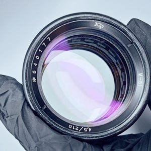May include: A black camera lens with a silver ring and a purple lens. The lens has numbers printed on the side, including "N 6 4 0 1 7 7" and "4.5/210".