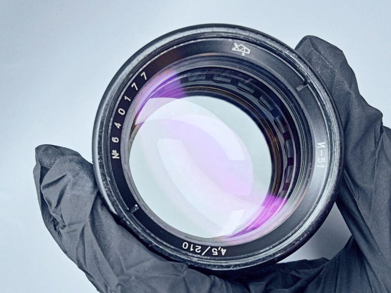May include: A black camera lens with a silver ring and a purple lens. The lens has numbers printed on the side, including "N 6 4 0 1 7 7" and "4.5/210".