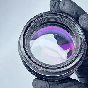 May include: A close-up of a black camera lens with a purple and blue colored glass element. The lens is being held by a gloved hand.