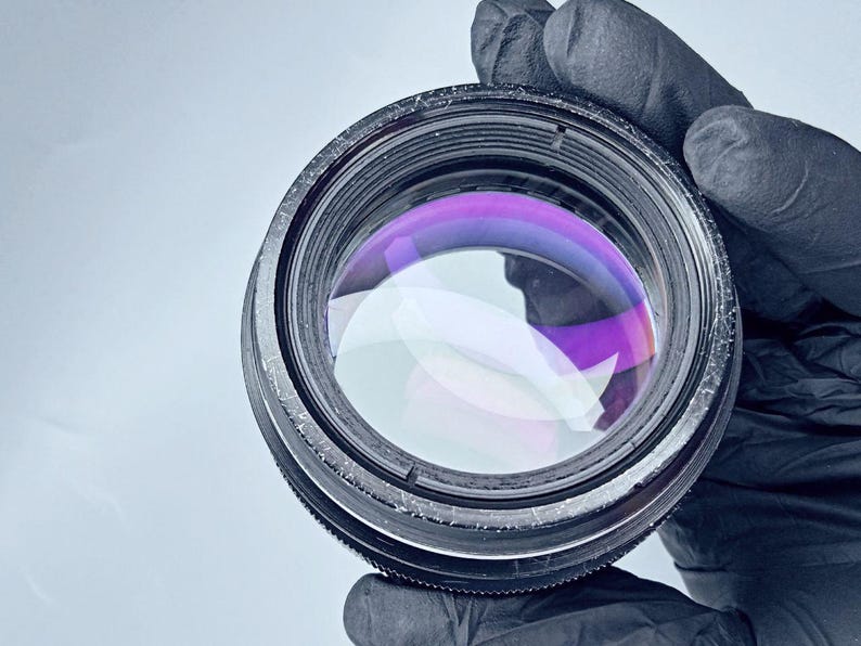 May include: A close-up of a black camera lens with a purple and blue colored glass element. The lens is being held by a gloved hand.