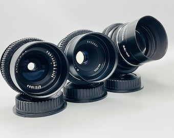 May include: Four black camera lenses with silver accents. The lenses are all different sizes and have different markings. The lenses are sitting on a white surface.