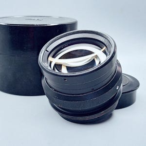 May include: A black camera lens with a silver ring and a white lens inside. The lens is in a black case.