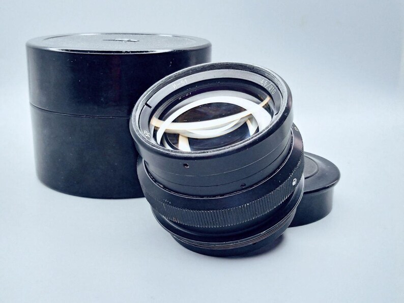 May include: A black camera lens with a silver ring and a white lens inside. The lens is in a black case.