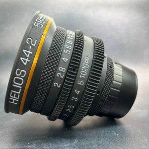 Puede incluir: Un objetivo Helios 44-2 de 58 mm negro con un anillo dorado alrededor del barril. El objetivo tiene una serie de números y marcas en el barril, incluyendo "2.8 4 5.6 8 11" y "6 10 20 ∞".