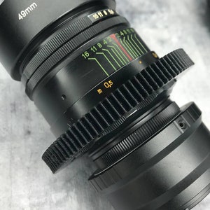 Könnte beinhalten: Ein schwarzes Kameraobjektiv mit einem 49 mm Filtergewinde und einer Tiefenschärfenskala, die in Metern gekennzeichnet ist. Das Objektiv hat einen Zahnkranz zum Fokussieren.
