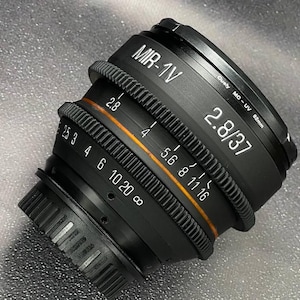 May include: Black MIR-1V 2.8/37 camera lens with white and orange markings. The lens has a ribbed focus ring and aperture settings from 2.8 to 16. The lens also has the text "MO-UV 52mm".