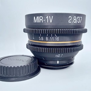 May include: A black camera lens with the text "MIR-1V 2.8/37" printed on the front. The lens has a gold ring with numbers 4, 5.6, 8, 11, and 16 printed on it. The lens also has a black lens cap.